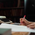 Hand mit rotem Stift über Manuskriptseiten neben einer Schreibmaschine