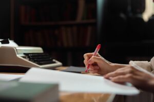 Hand mit rotem Stift über Manuskriptseiten neben einer Schreibmaschine