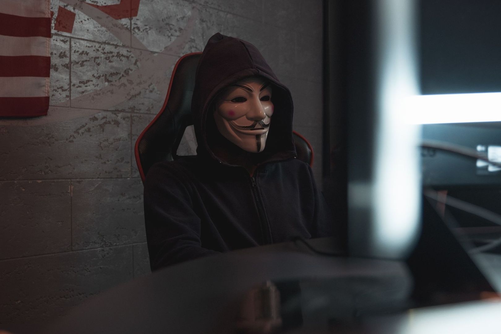Anonym wirkende Person mit Guy-Fawkes-Maske sitzt vor einem Monitor
